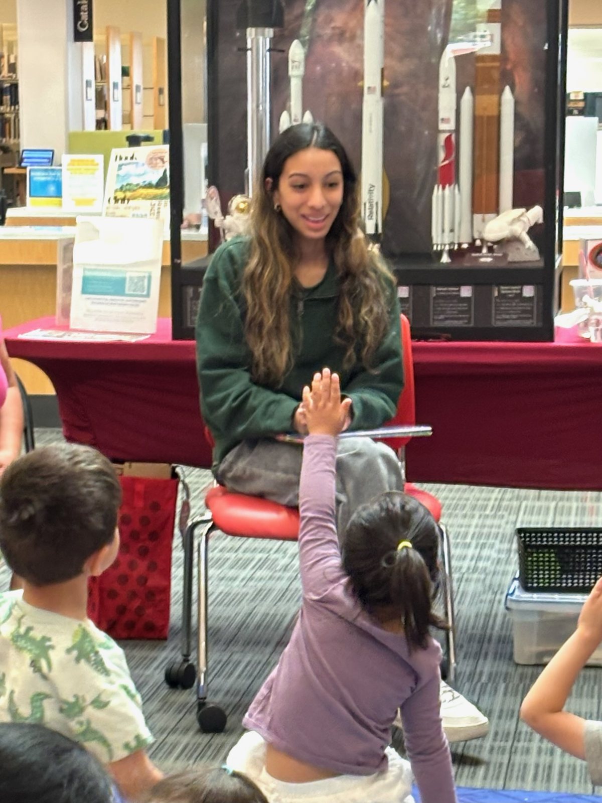 A young child raising a hand high during storytime as Saanvi Phaneendra smiles in front of the rocket display