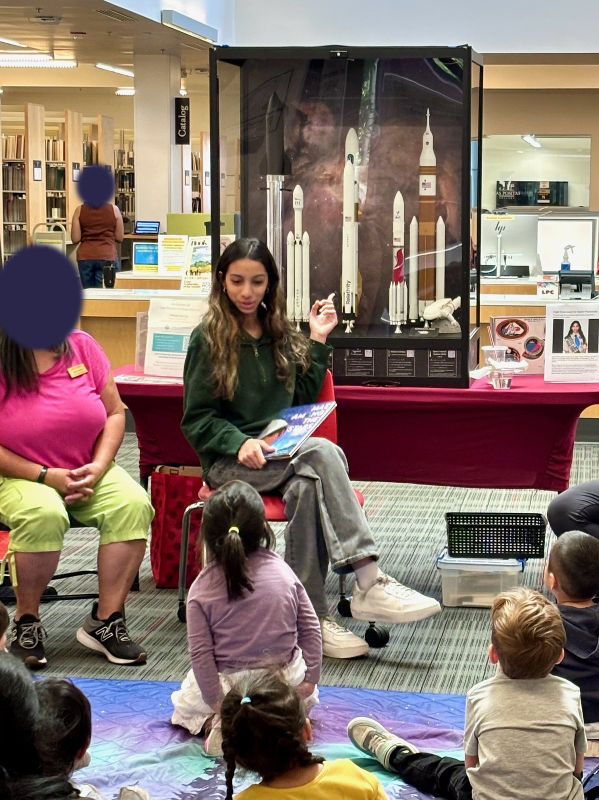Wide view of storytime at the Las Positas College Library, with Saanvi Phaneendra reading in front of the Galactic Getaway rocket exhibit and children seated on a mat