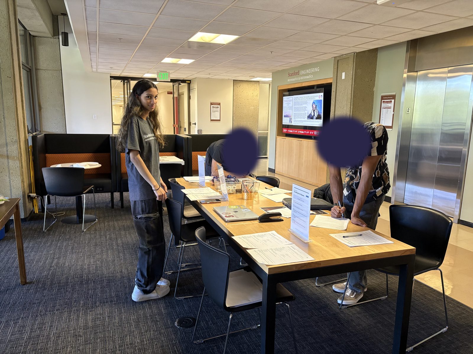 Saanvi standing at the survey table while two participants fill out forms. Participant faces are covered.