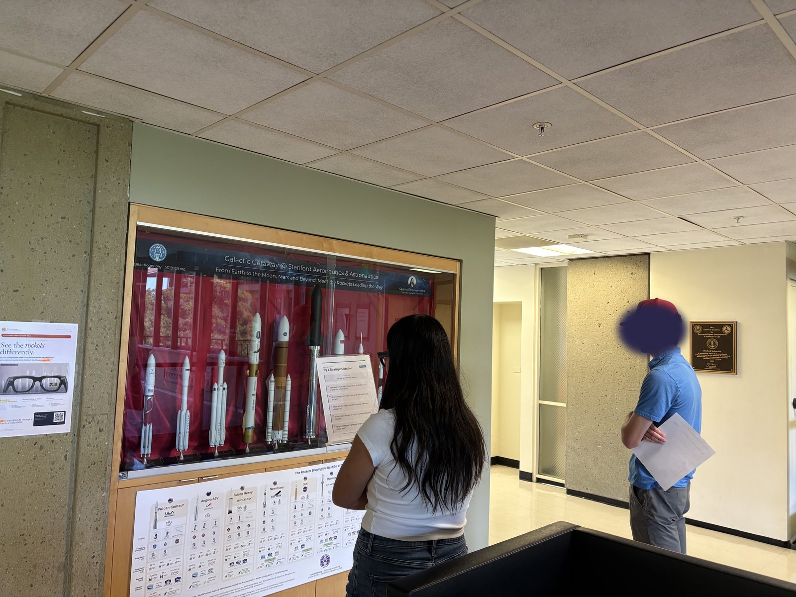 Two participants looking at the rocket display case and comparison poster. Faces are covered.