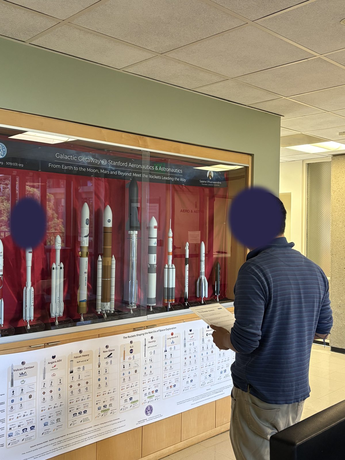 A participant holding a question card in front of the rocket display case. Face is covered.